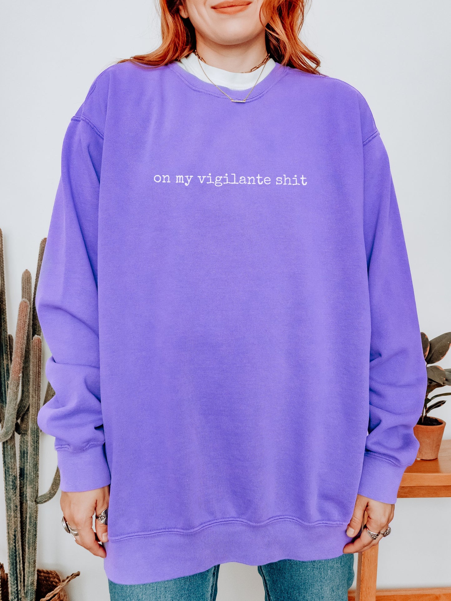Person wearing a purple sweatshirt with text, standing indoors with plants in the background