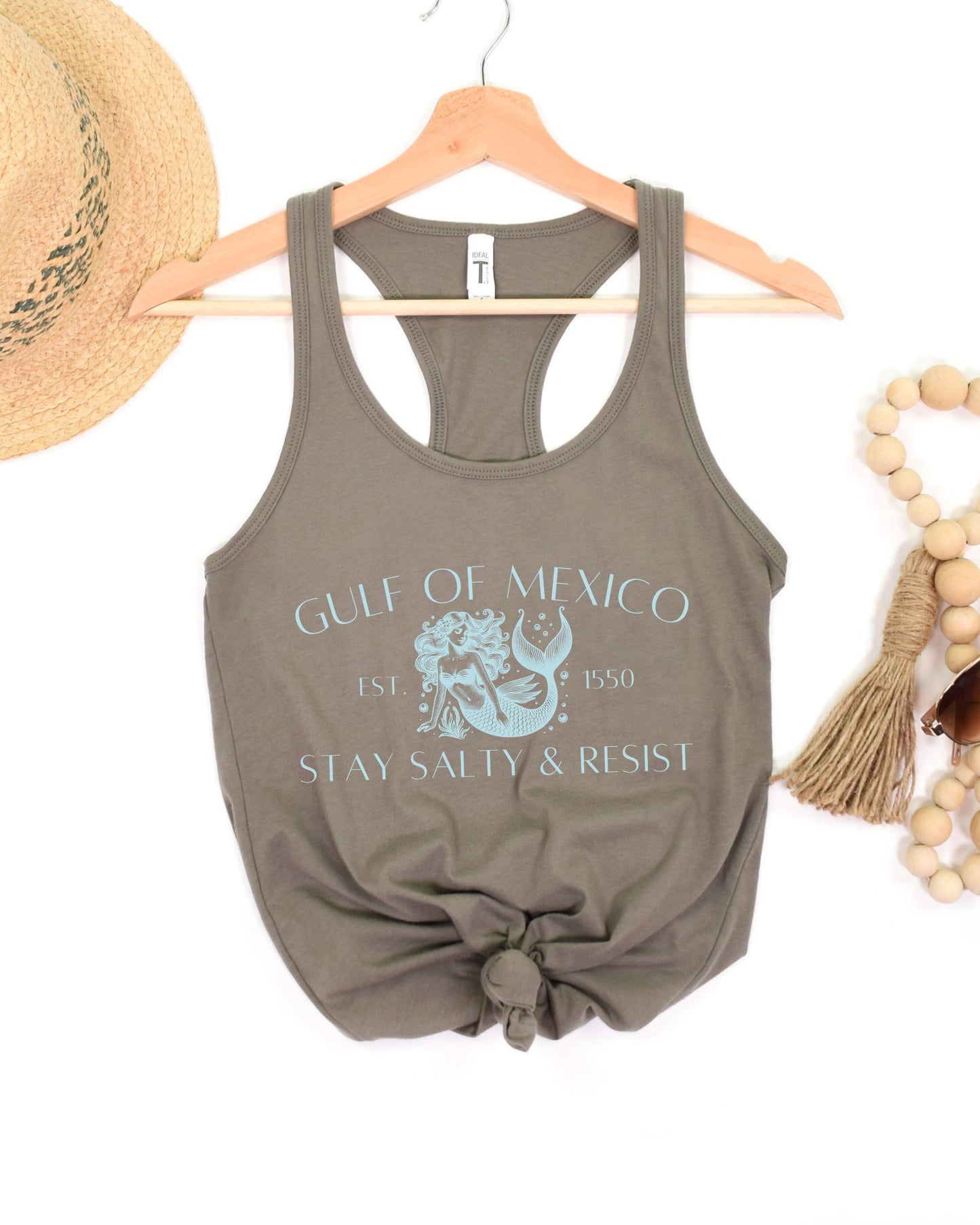 Olive green tank top with graphic design and text on a hanger, surrounded by a hat and beads.