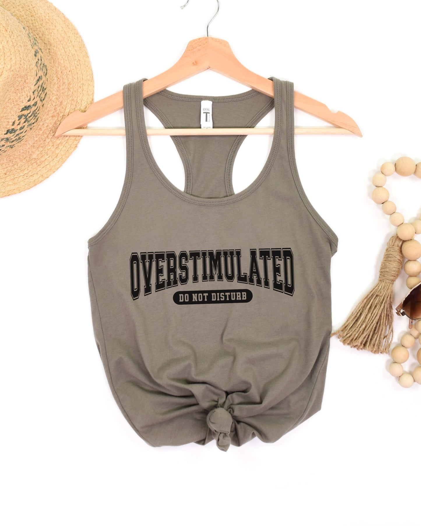 Gray tank top with 'OVERSTIMULATED DO NOT DISTURB' text on a hanger with accessories.