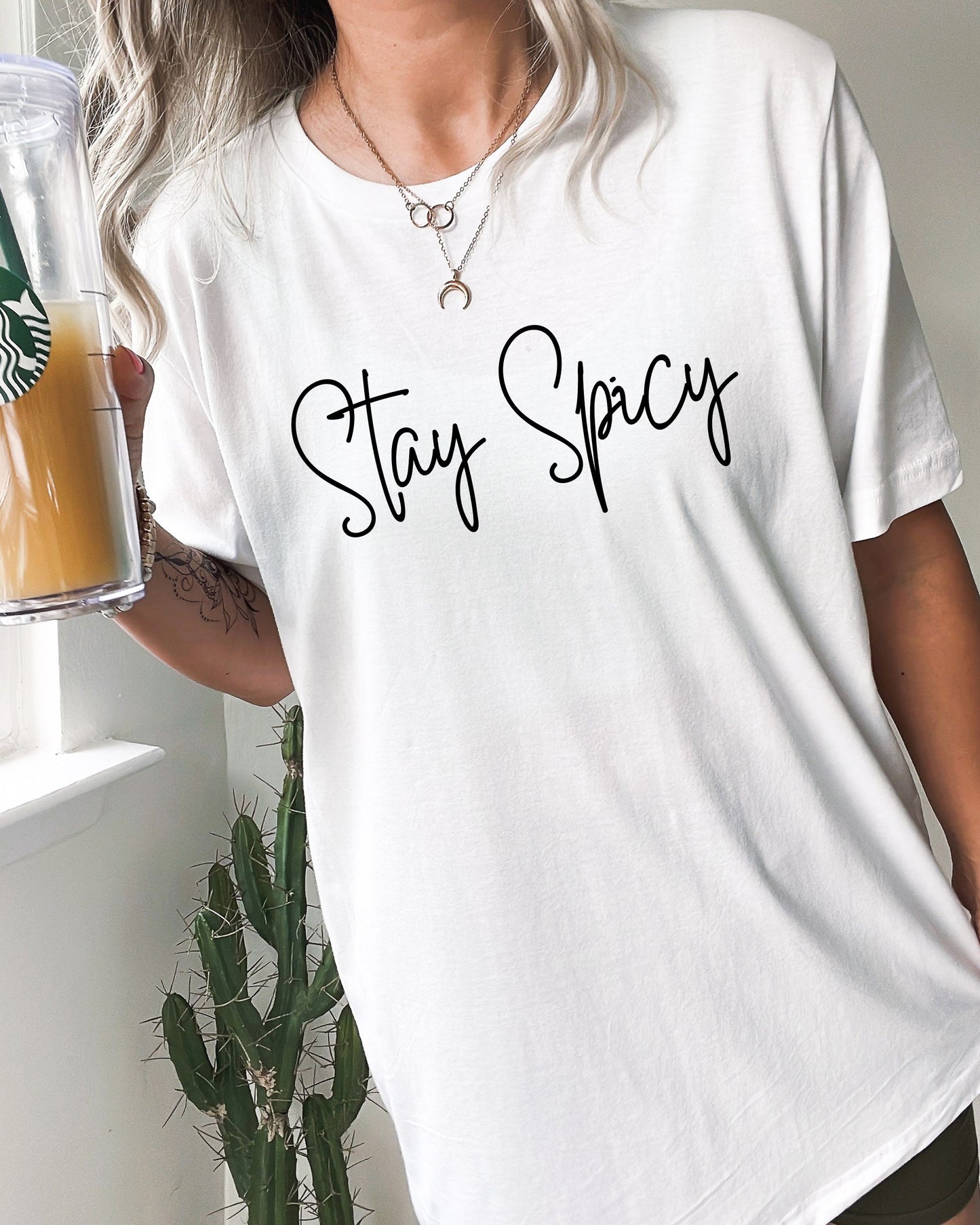 Person wearing a white t-shirt with 'Stay Spicy' text, holding a Starbucks cup.
