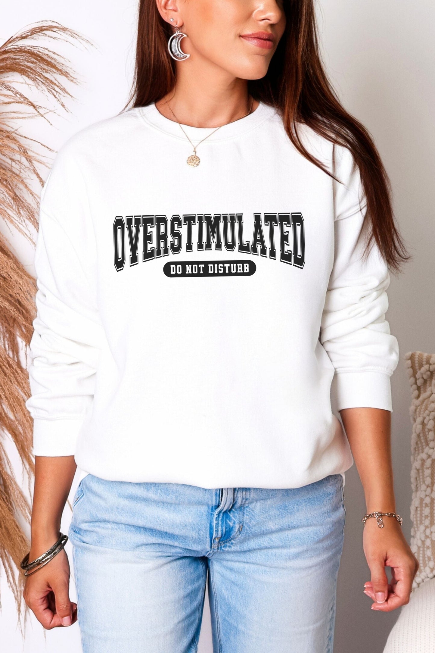 Person wearing a white sweatshirt with 'OVERSTIMULATED DO NOT DISTURB' text, standing against a neutral background.