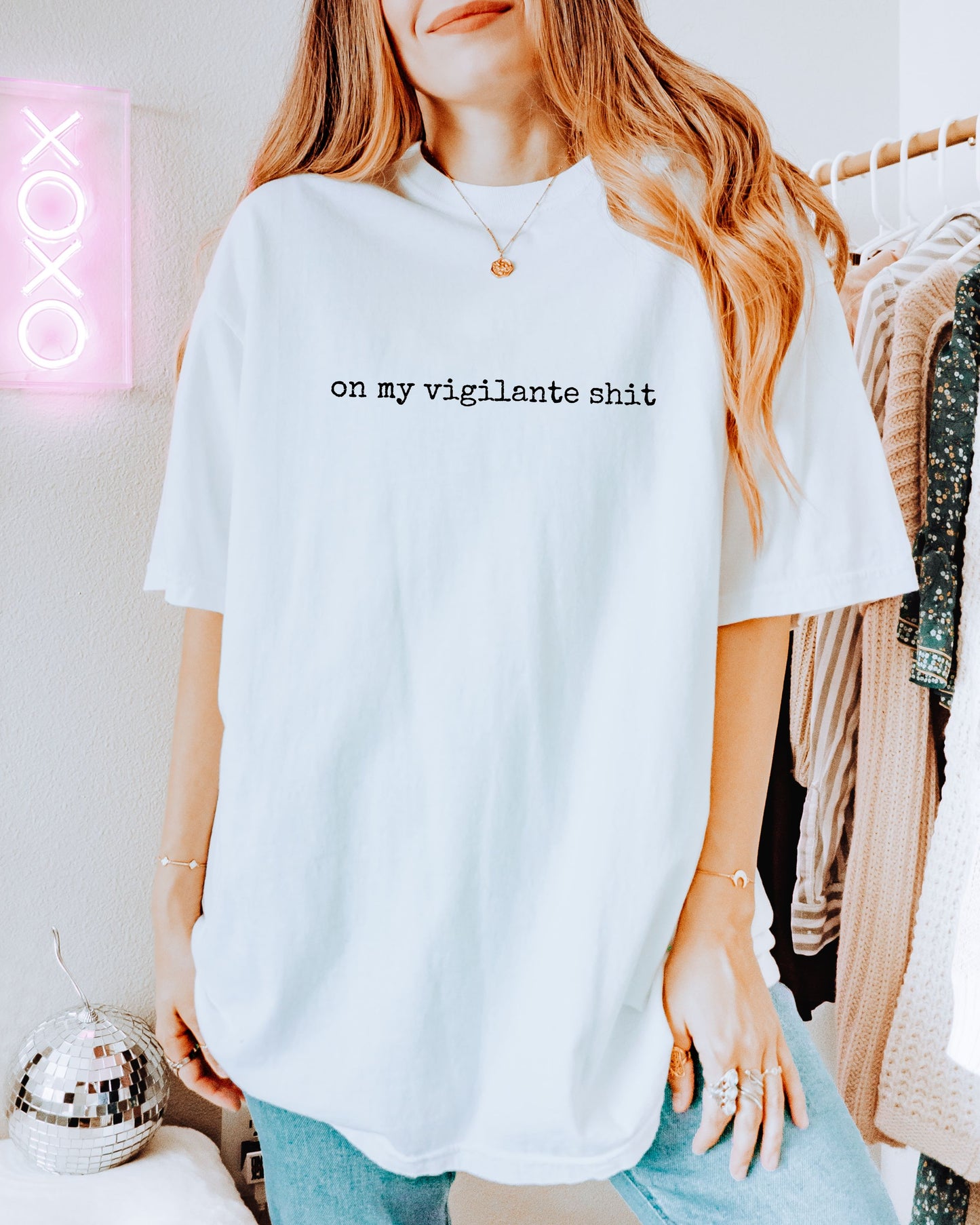 Person wearing a white t-shirt with on my vigilante shit text in black font, standing in a room with a neon sign and clothing rack.
