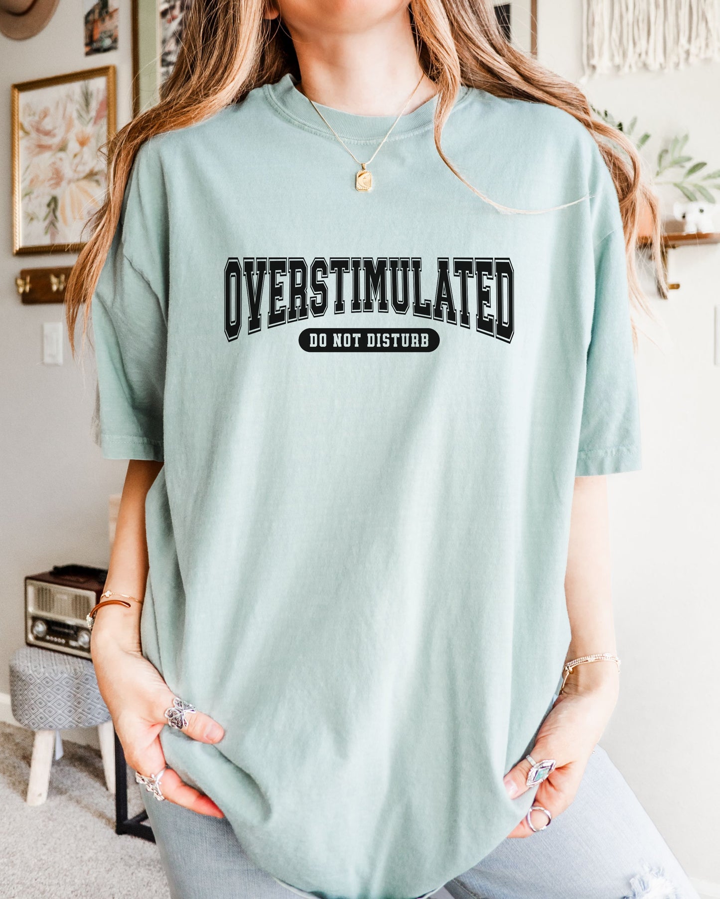 Person wearing a light blue t-shirt with 'OVERSTIMULATED DO NOT DISTurb' text indoors.