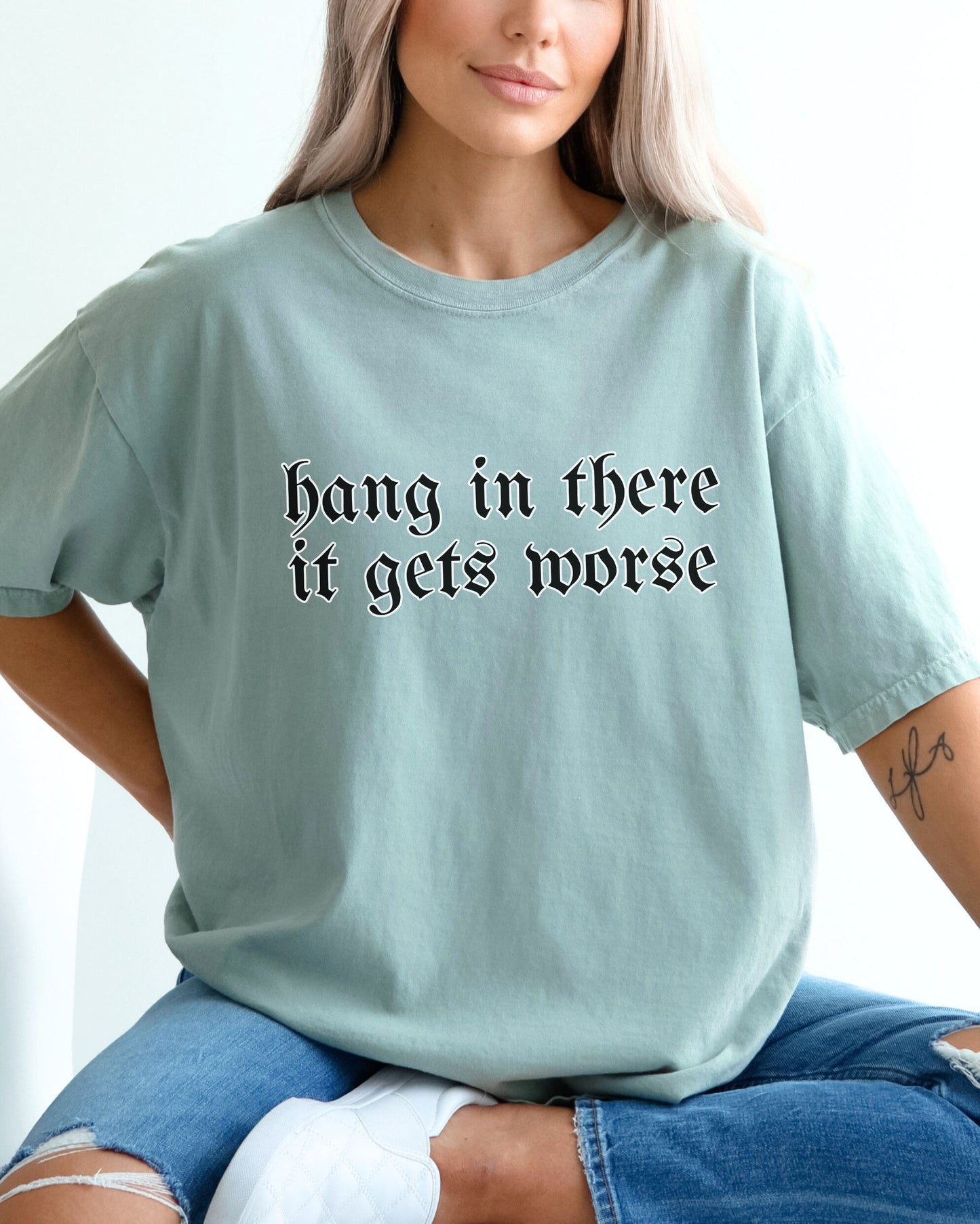 Person wearing a light blue t-shirt with text 'hang in there it gets worse' on a plain background