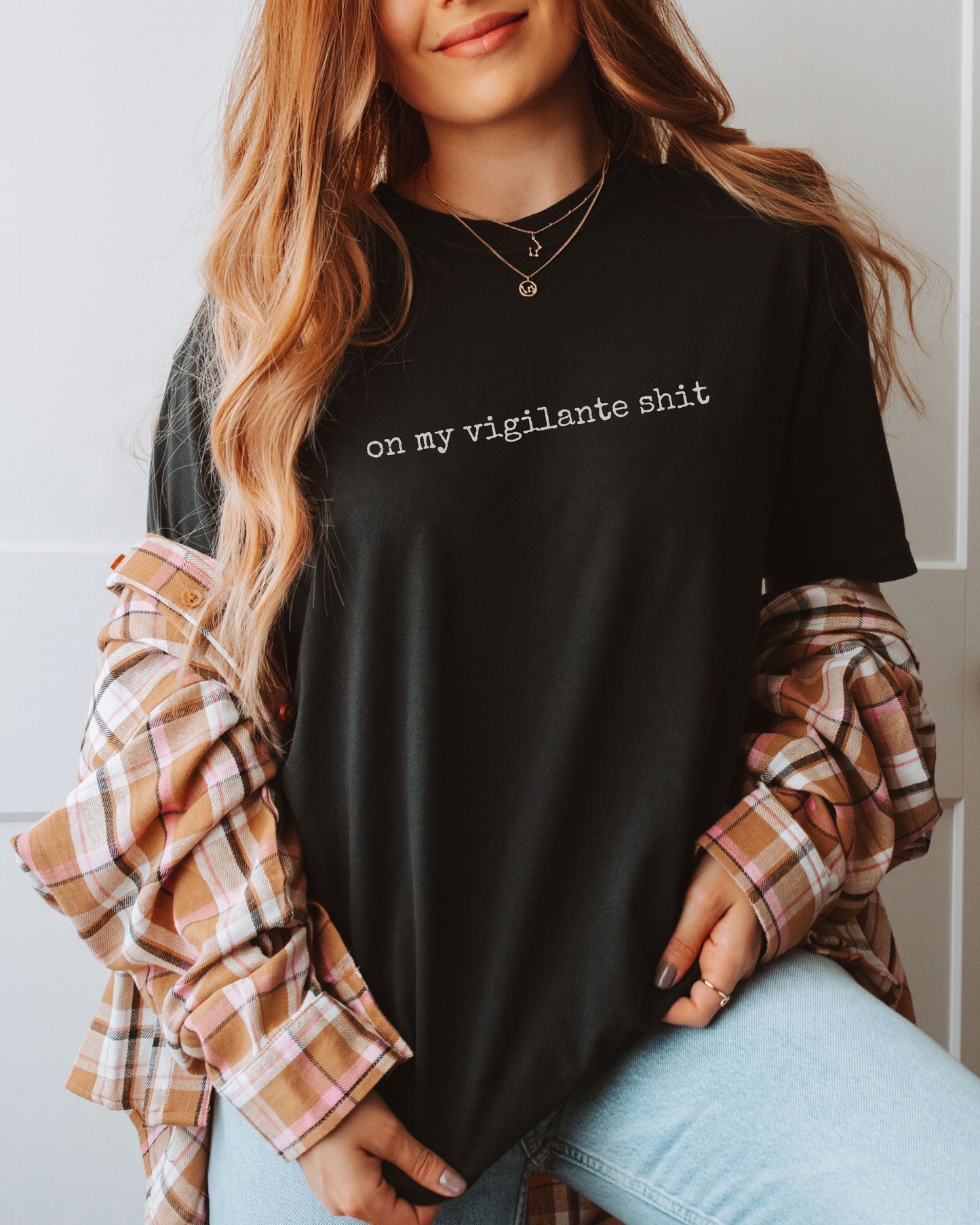 Person wearing a black t-shirt with on my vigilante shit text in white font, plaid sleeves, and light blue jeans.