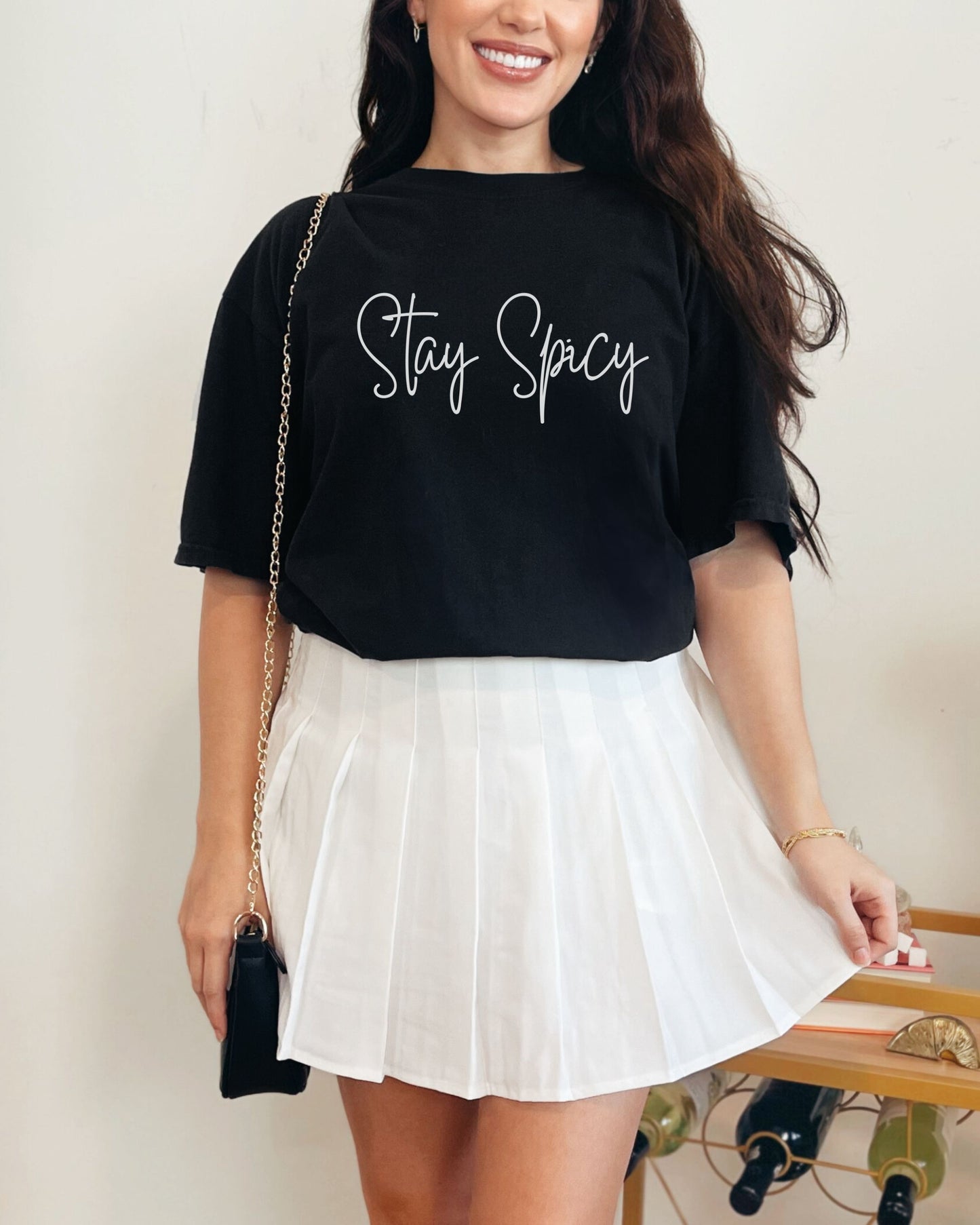 Woman wearing a black t-shirt with 'Stay Spicy' text and a white skirt, standing against a plain background.