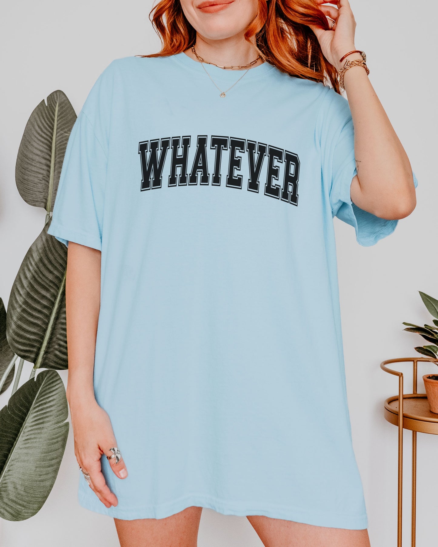 Person wearing a light blue t-shirt with 'WHATEVER' text, standing indoors with plants in the background.