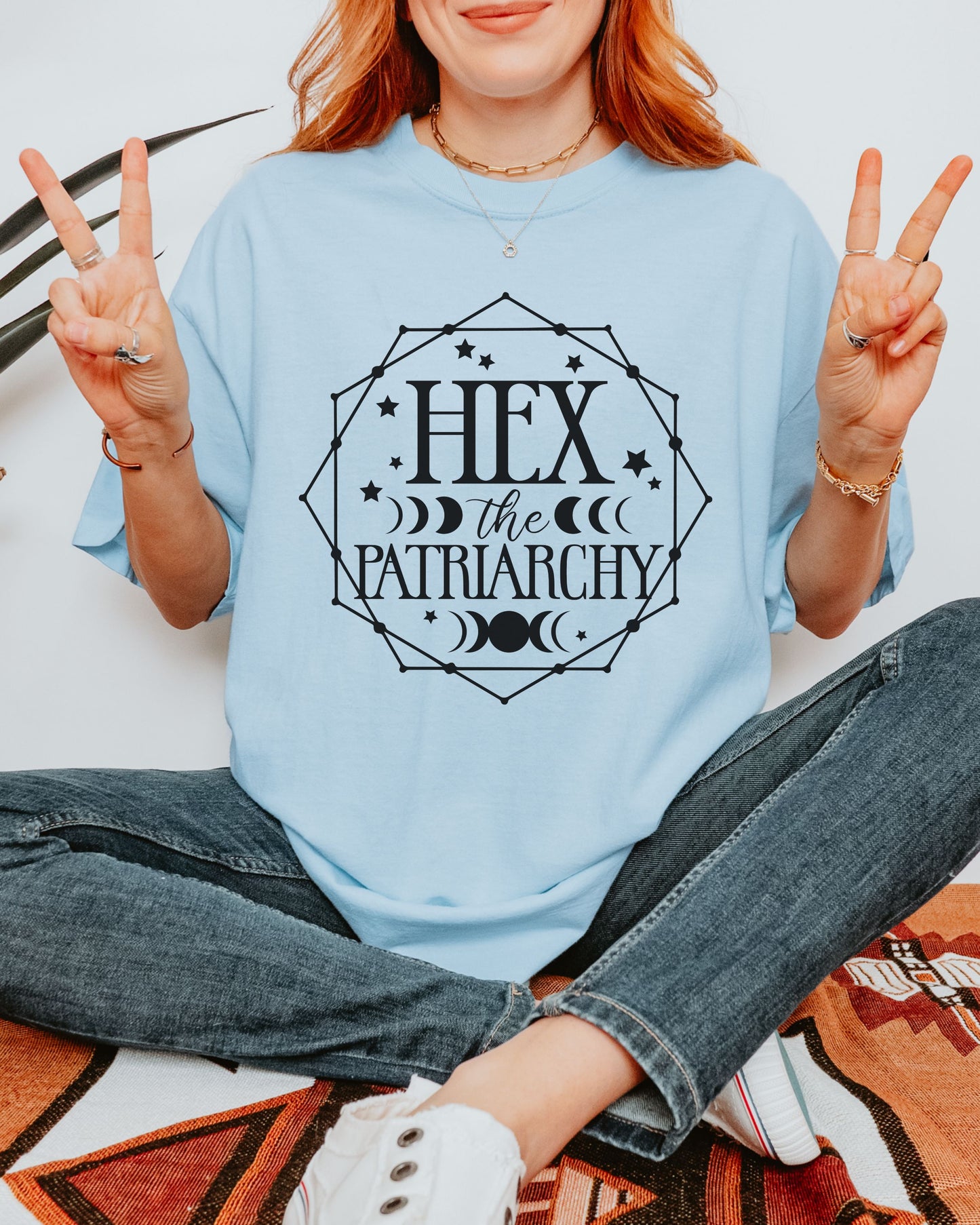 Person wearing a light blue shirt with 'Hex the Patriarchy' text, sitting on a patterned rug.