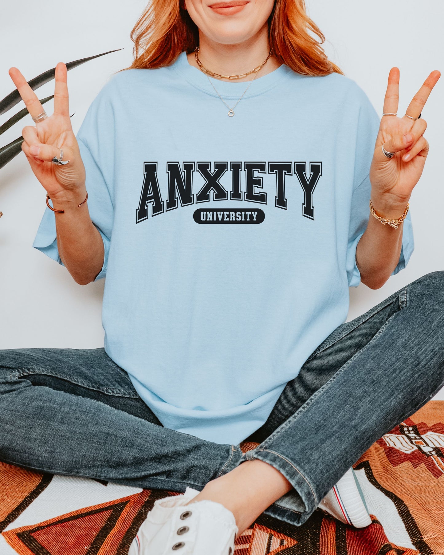 Person wearing a light blue shirt with 'ANXIETY UNIVERSITY' text, sitting on a patterned rug.