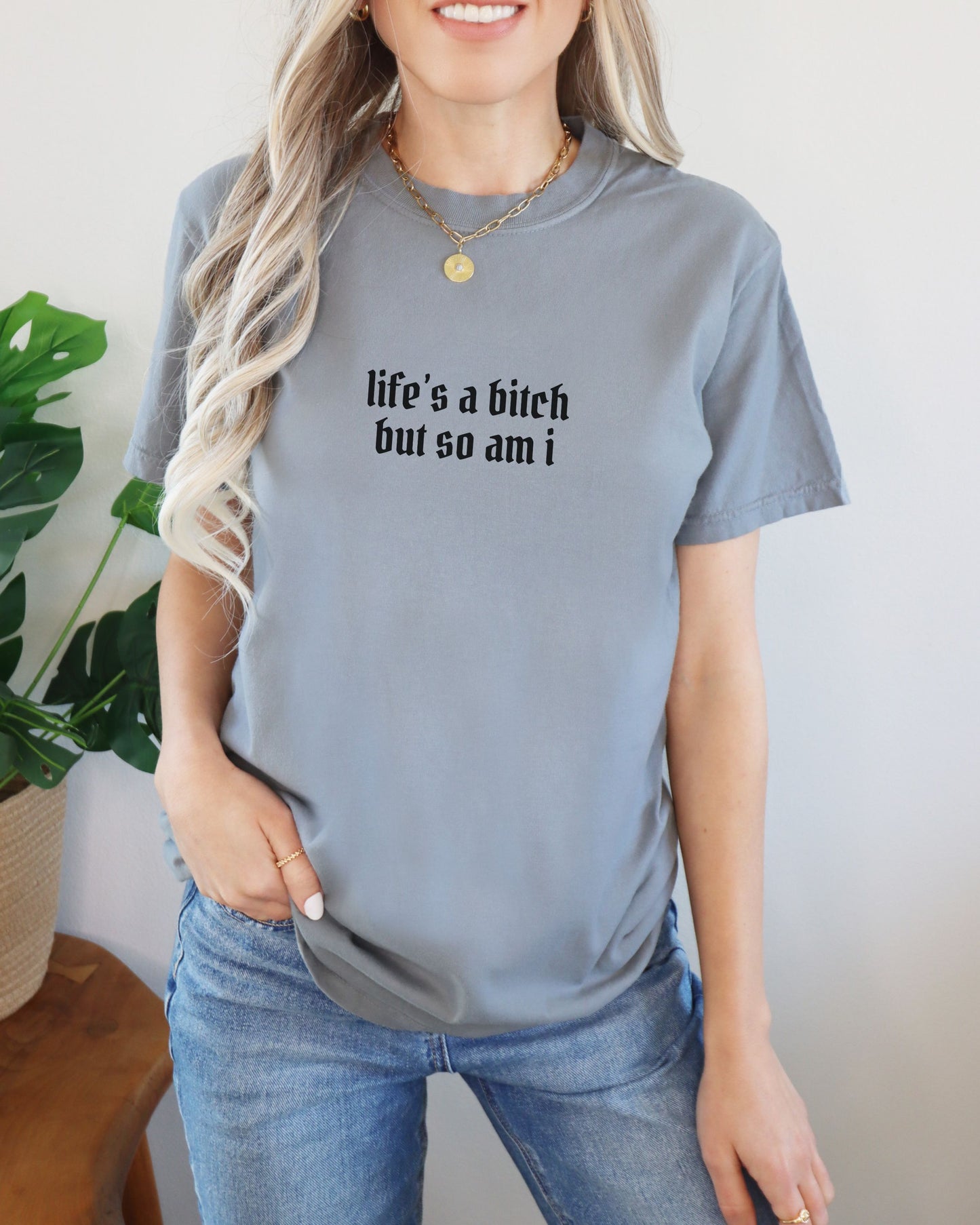 Person wearing a gray t-shirt with text, standing indoors with a plant in the background