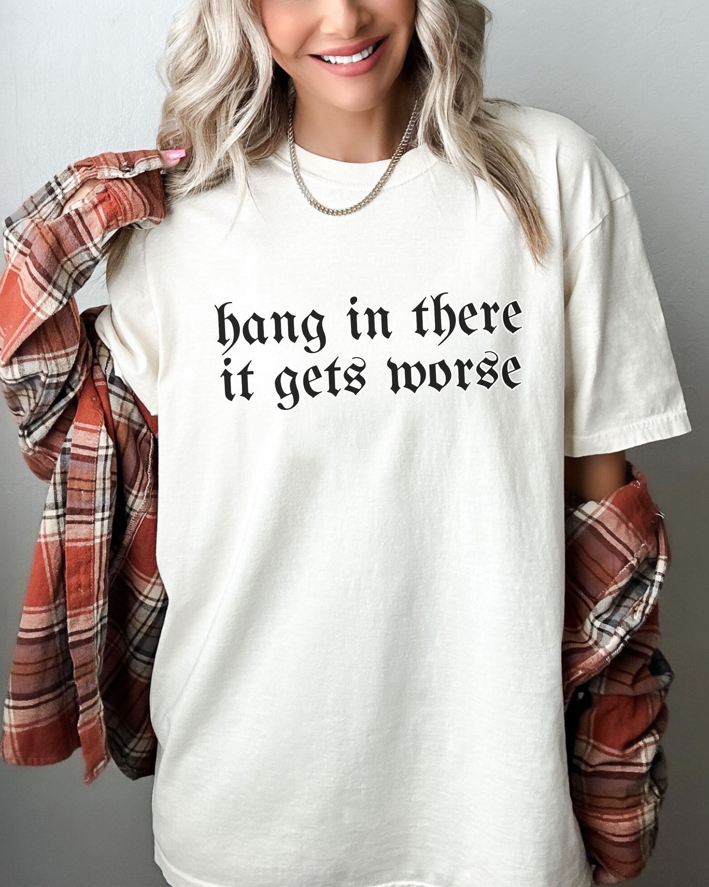 Person wearing a white t-shirt with 'hang in there it gets worse' text, holding a plaid shirt.