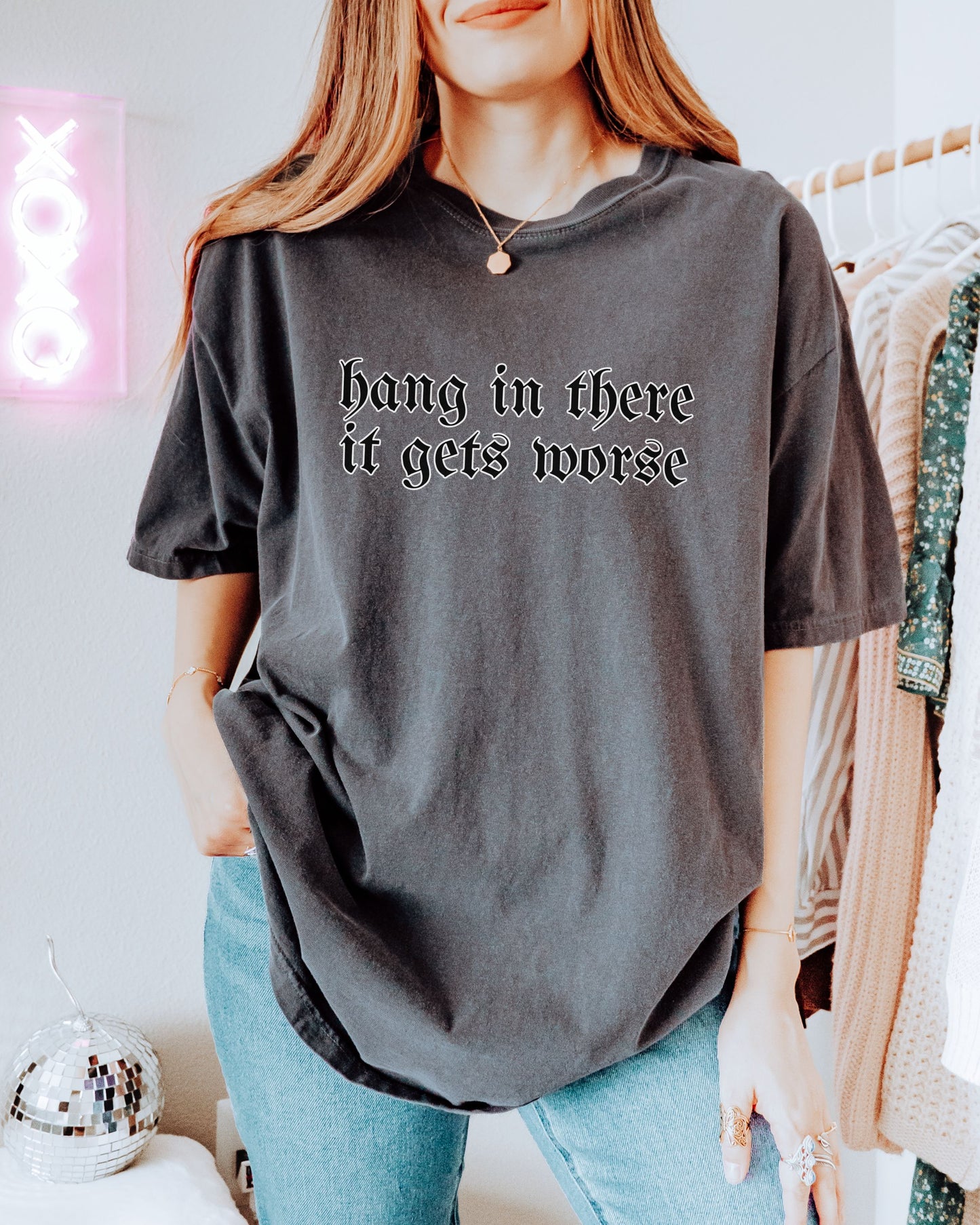 Person wearing a dark gray t-shirt with text, standing in a room with a neon sign and clothing rack.