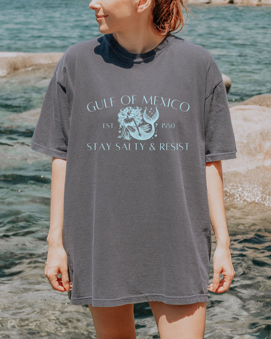 Person wearing a gray t-shirt with 'Gulf of Mexico' design by the water