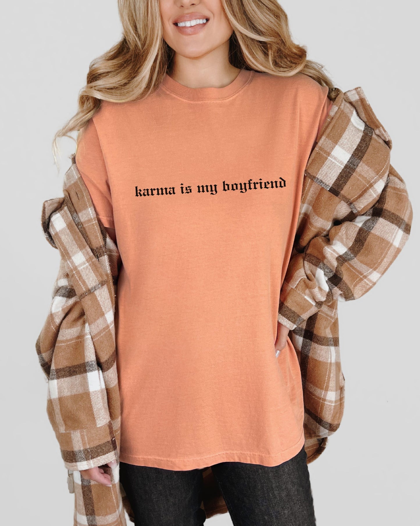 Person wearing an orange t-shirt with 'karma is my boyfriend' text, paired with a plaid shirt over shoulders on a light gray background.