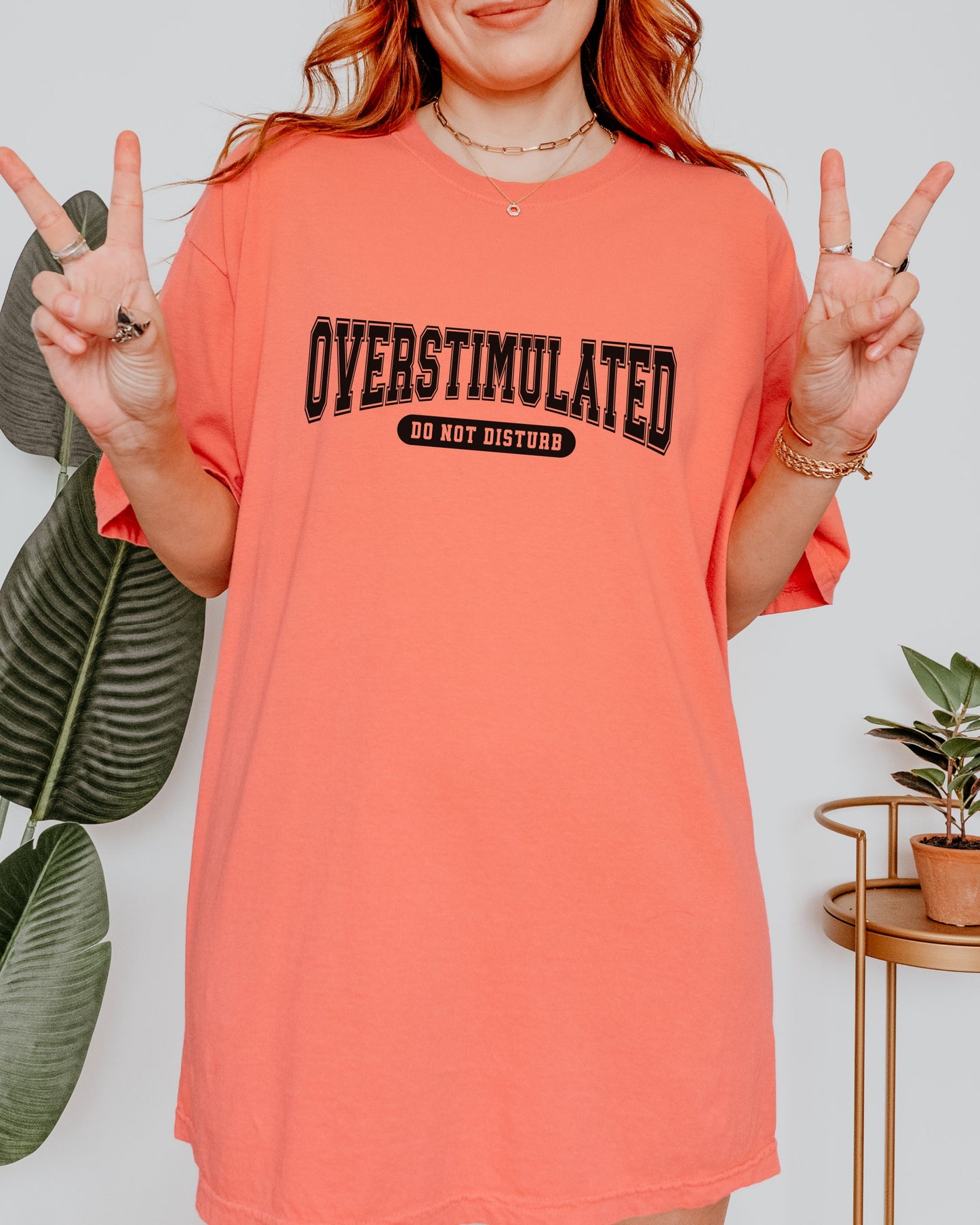 Person wearing a coral t-shirt with 'OVERSTIMULATED DO NOT DISTURB' text, making peace signs.