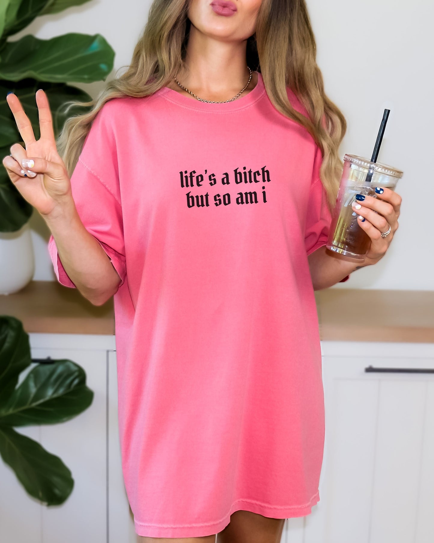 Person wearing a pink shirt with text, making a peace sign and holding a drink.