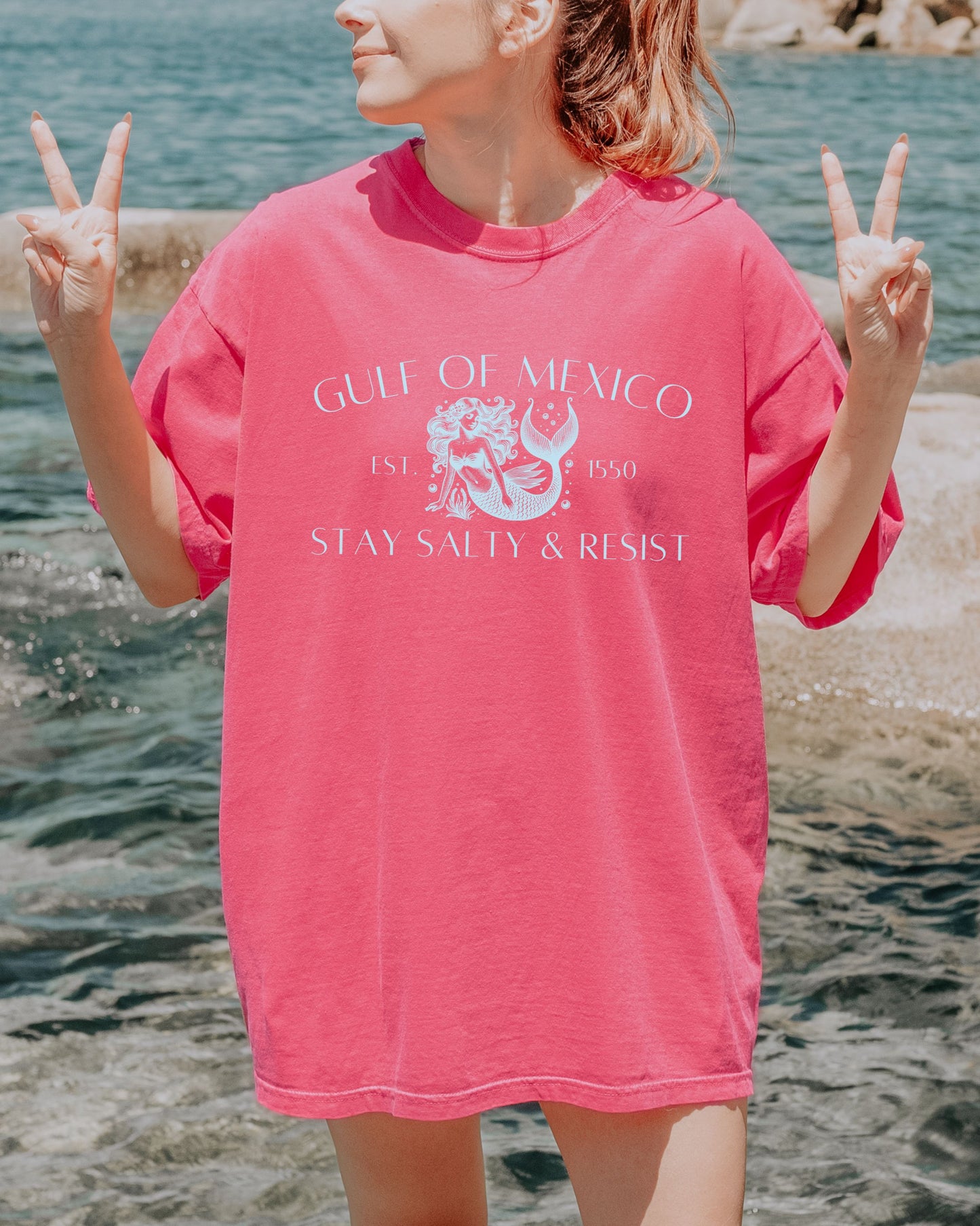 Person wearing a pink 'Gulf of Mexico' shirt by the water, making peace signs.