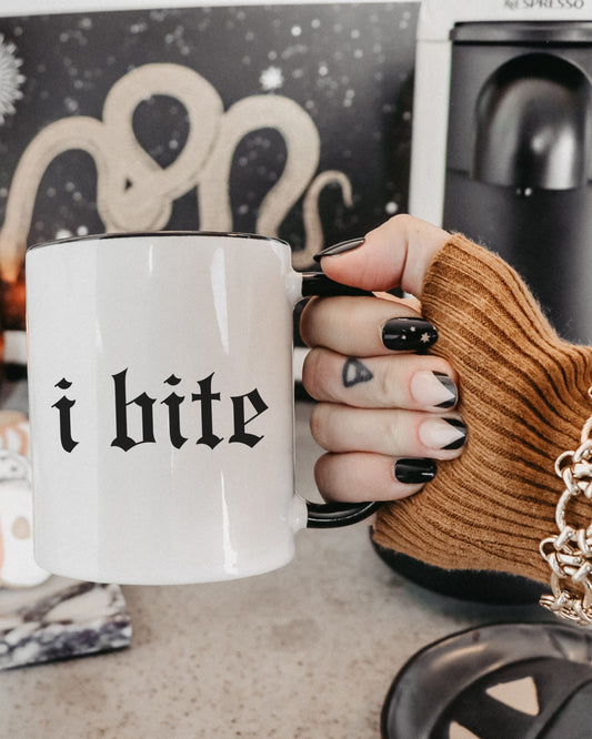 Hand holding a white mug with 'i bite' text, wearing a brown glove, against a blurred kitchen background.