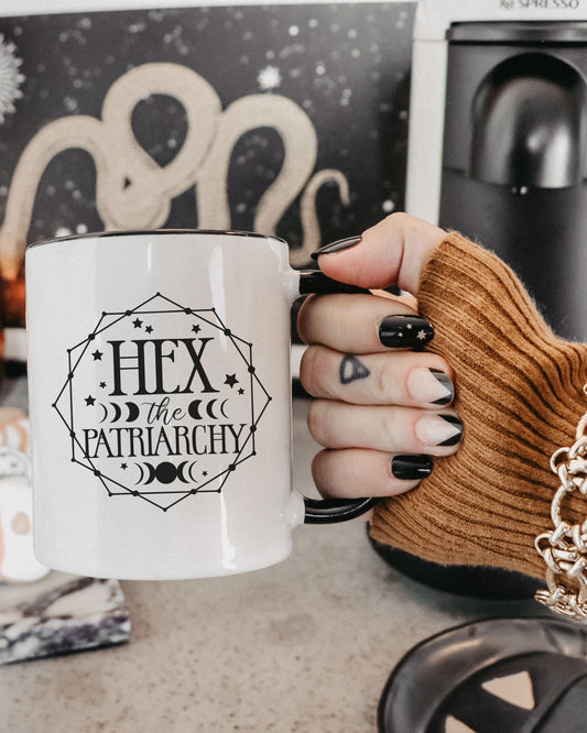 Hand holding a mug with 'Hex the Patriarchy' text in front of a coffee machine.
