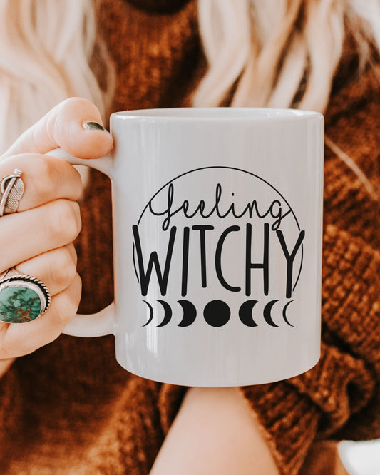 Person holding a white mug with 'Feeling Witchy' text and moon phases design.