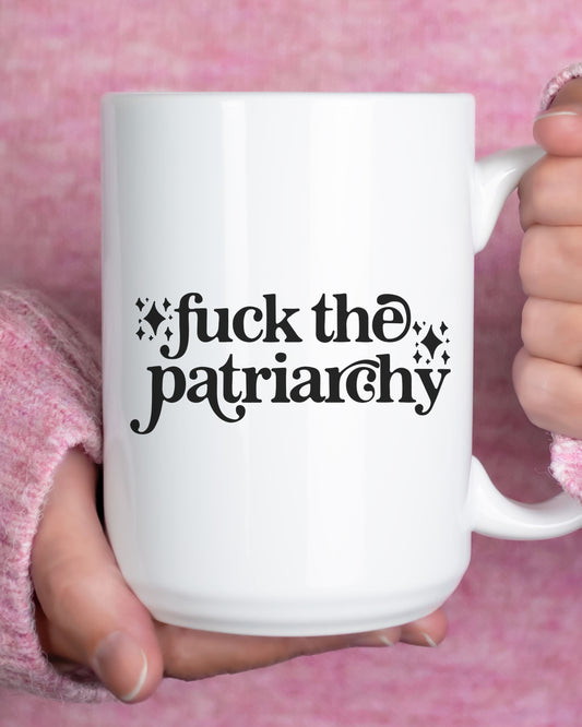 White mug with 'fuck the patriarchy' text held by a person against a pink background