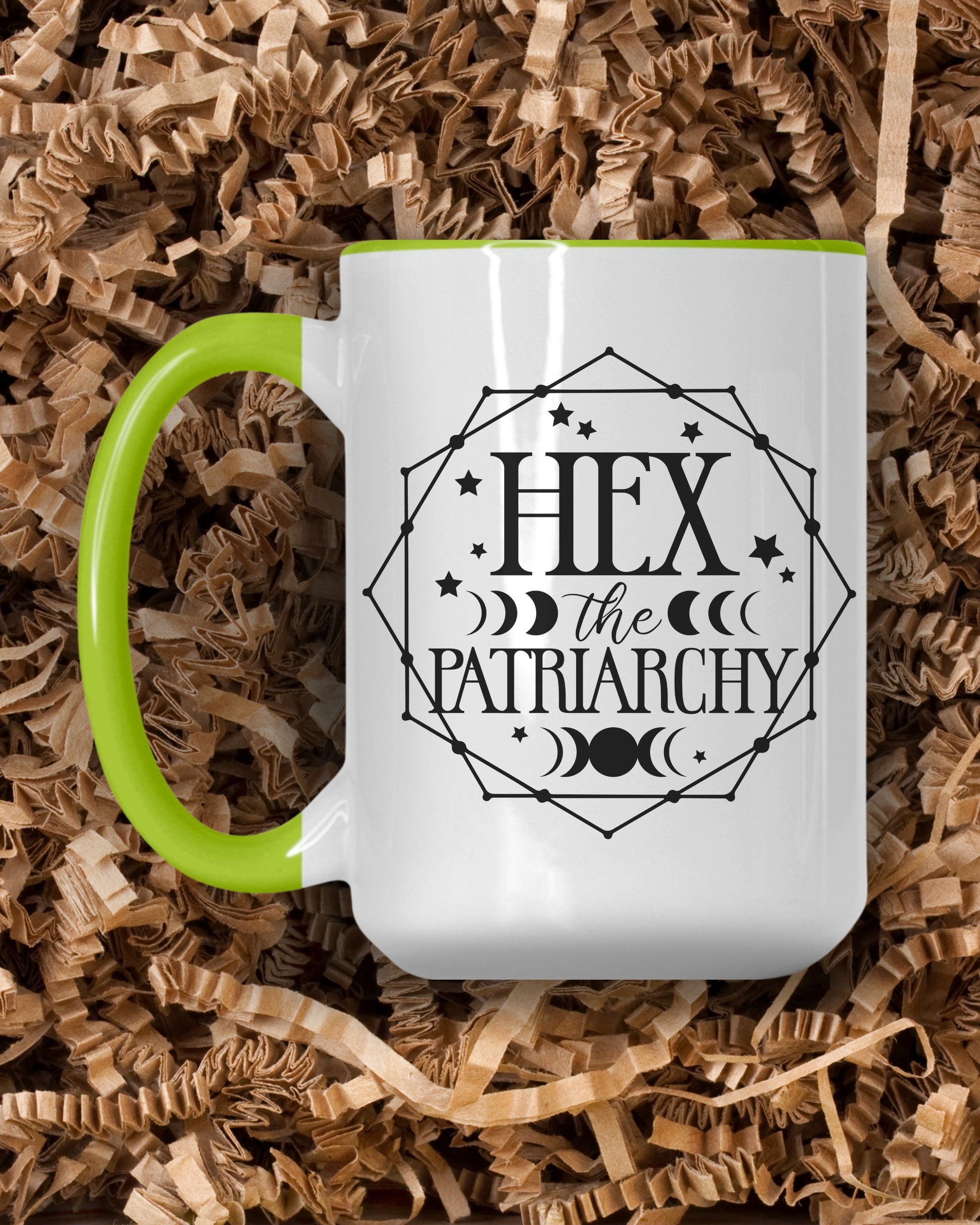 White mug with green handle featuring 'Hex the Patriarchy' design on a bed of brown shredded paper.