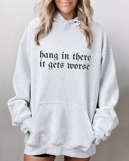 Gray hoodie with 'hang in there it gets worse' text worn by a person on a plain background