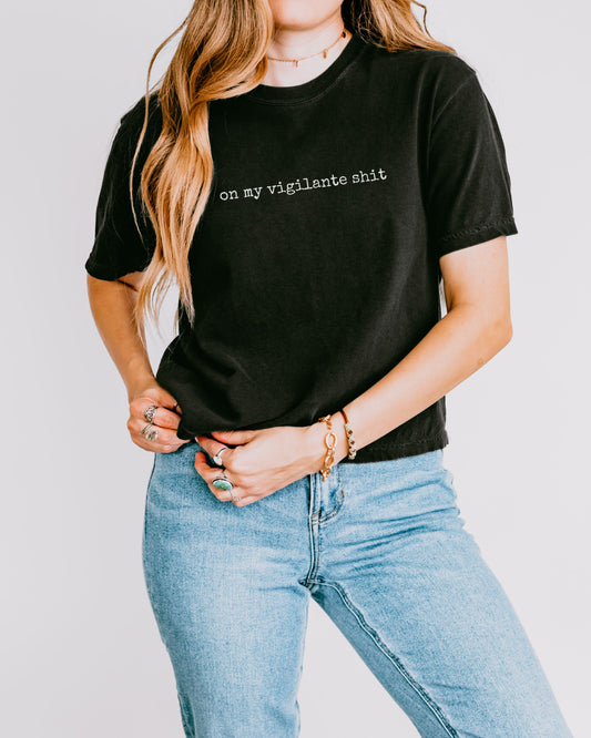 Person wearing a black t-shirt with on my vigilante shit text in white font  and blue jeans on a plain background