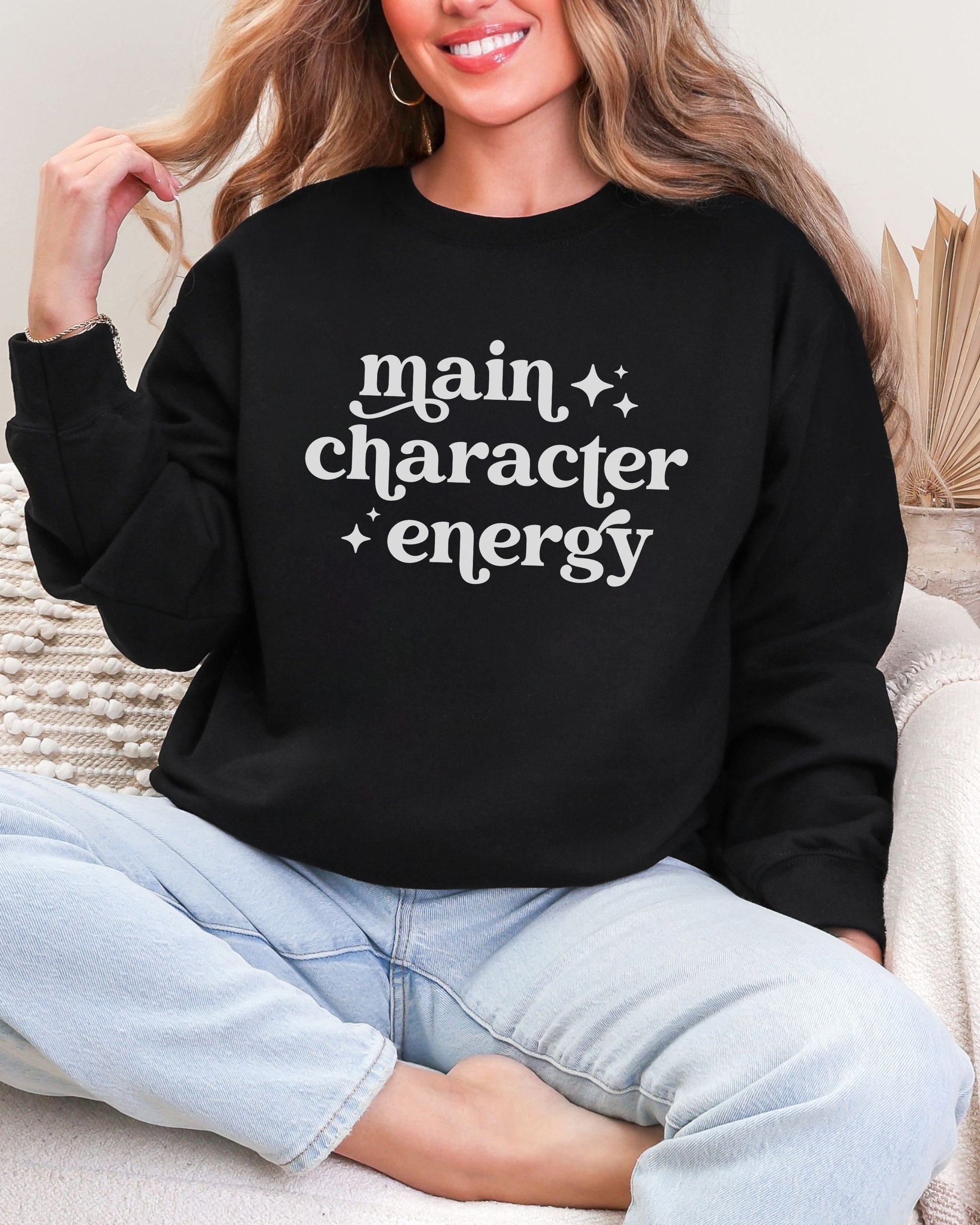 Person wearing a black sweatshirt with 'main character energy' text, sitting on a couch.
