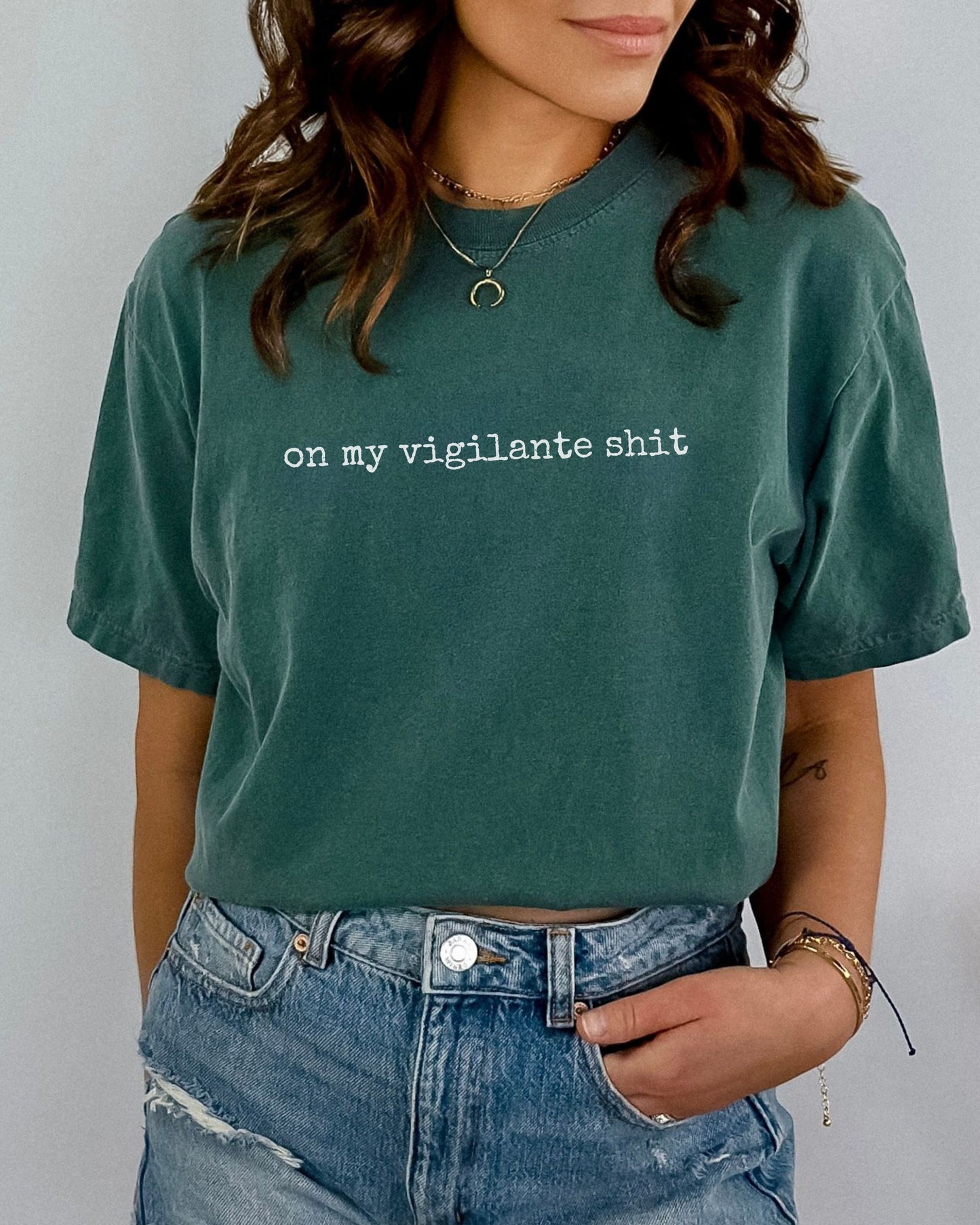 Person wearing a green t-shirt with on my vigilante shit text in white font and blue jeans against a plain background