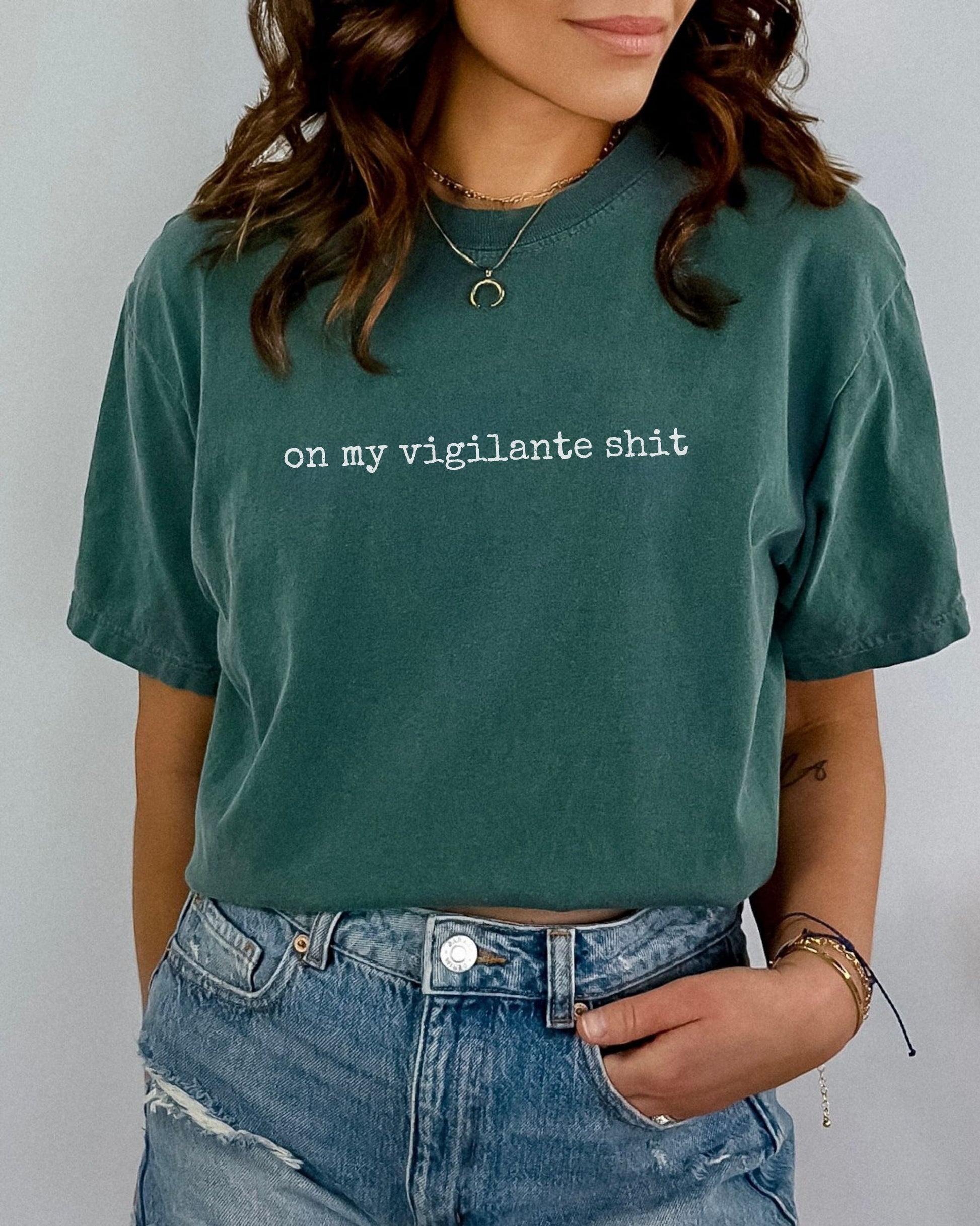 Person wearing a green t-shirt with on my vigilante shit text in white font and blue jeans against a plain background