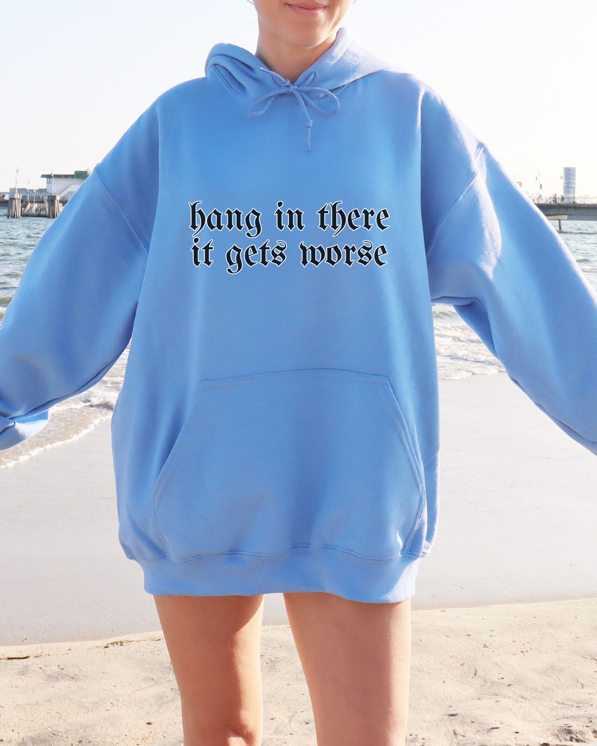 Person wearing a blue hoodie with text 'hang in there it gets worse' by a waterfront.