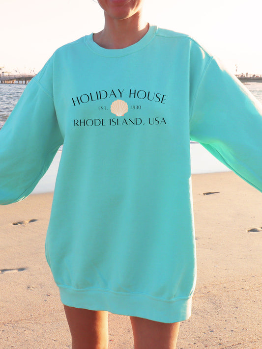 Person wearing a teal sweatshirt with 'Holiday House' text on a beach