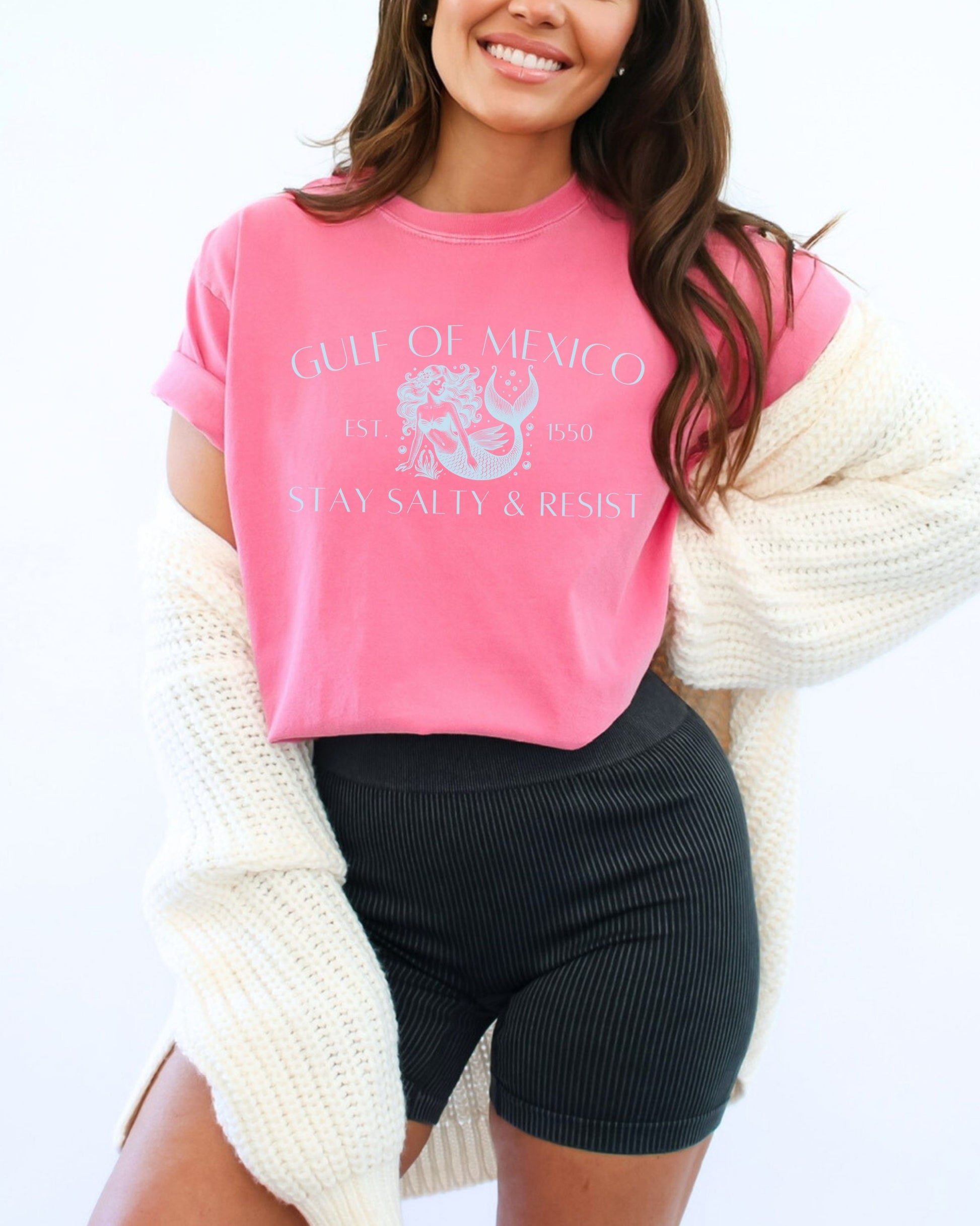 Woman wearing a pink graphic t-shirt with text and graphics, paired with black shorts and a white cardigan on a white background.