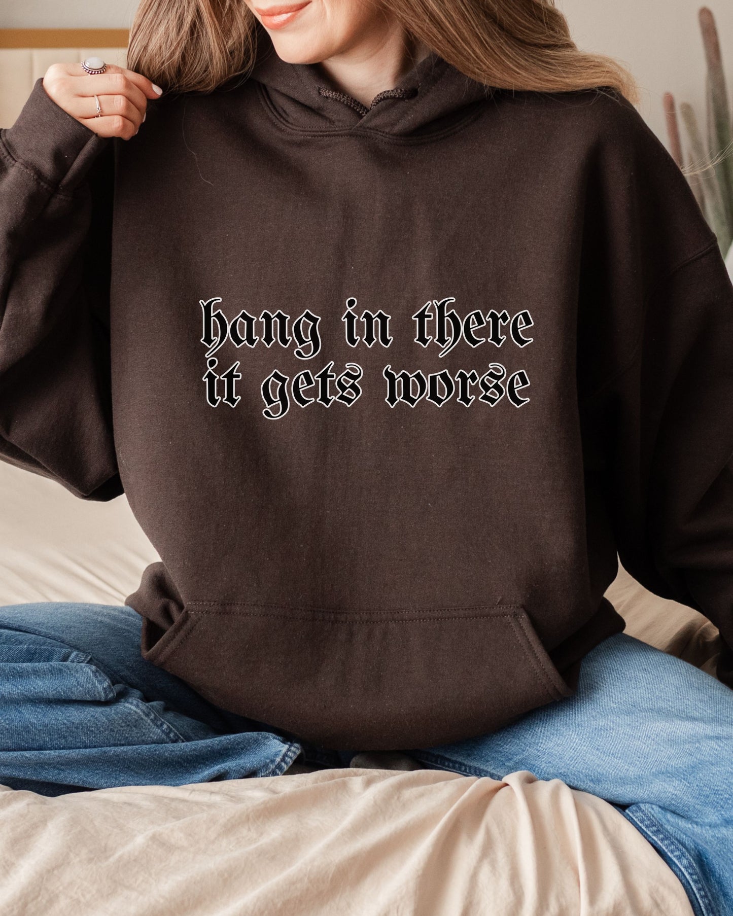 Person wearing a brown hoodie with text 'hang in there it gets worse' sitting on a couch.