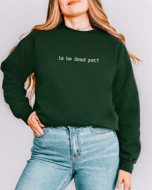 person wearing a forest green crewneck sweatshirt with "is he dead yet?" printed in a typewriter font standing in front of a white wall