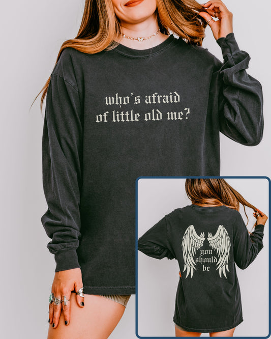 Black long-sleeve shirt with text 'who's afraid of little old me?' on front, and 'you should be' with wings on back, worn by a person.
