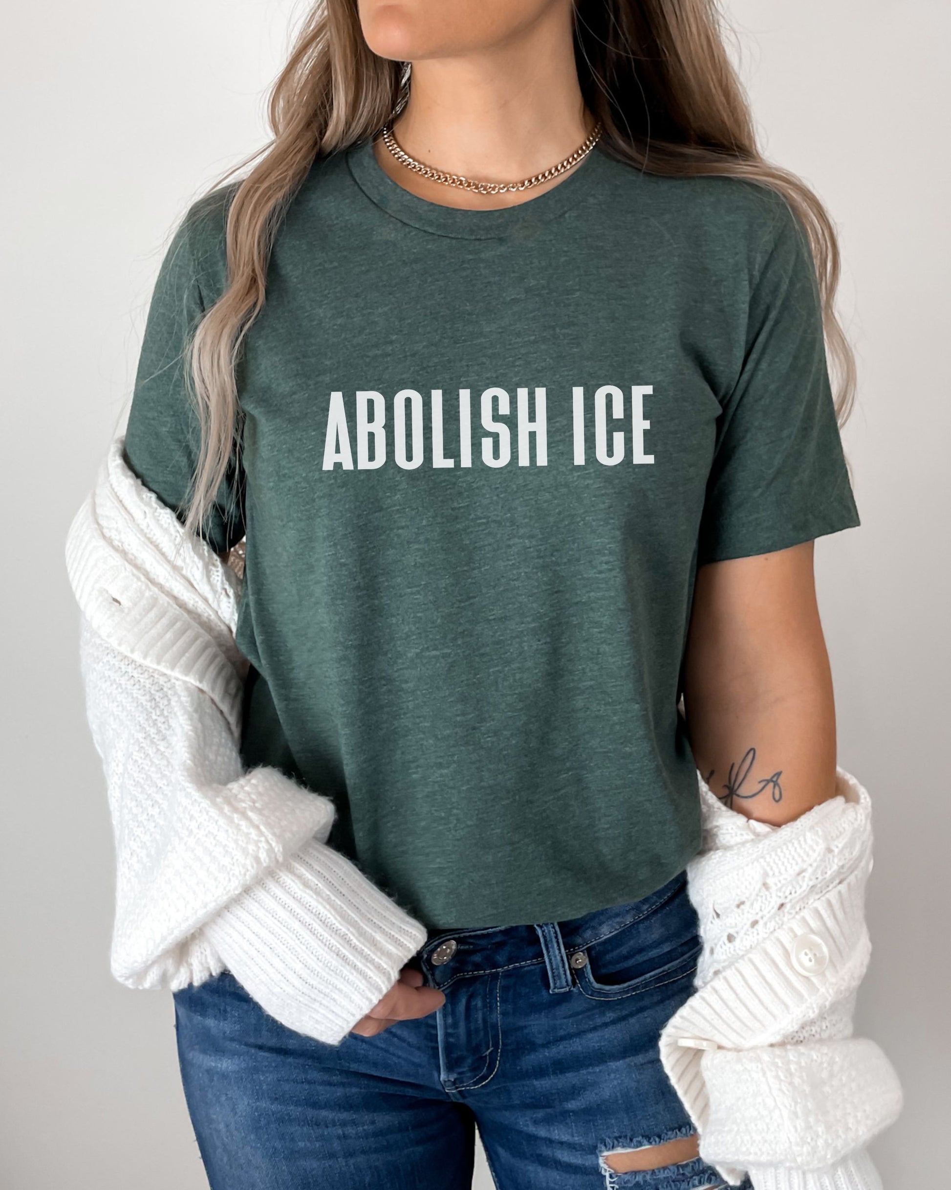 Person wearing a green t-shirt with 'ABOLISH ICE' text, white sweater, and blue jeans.