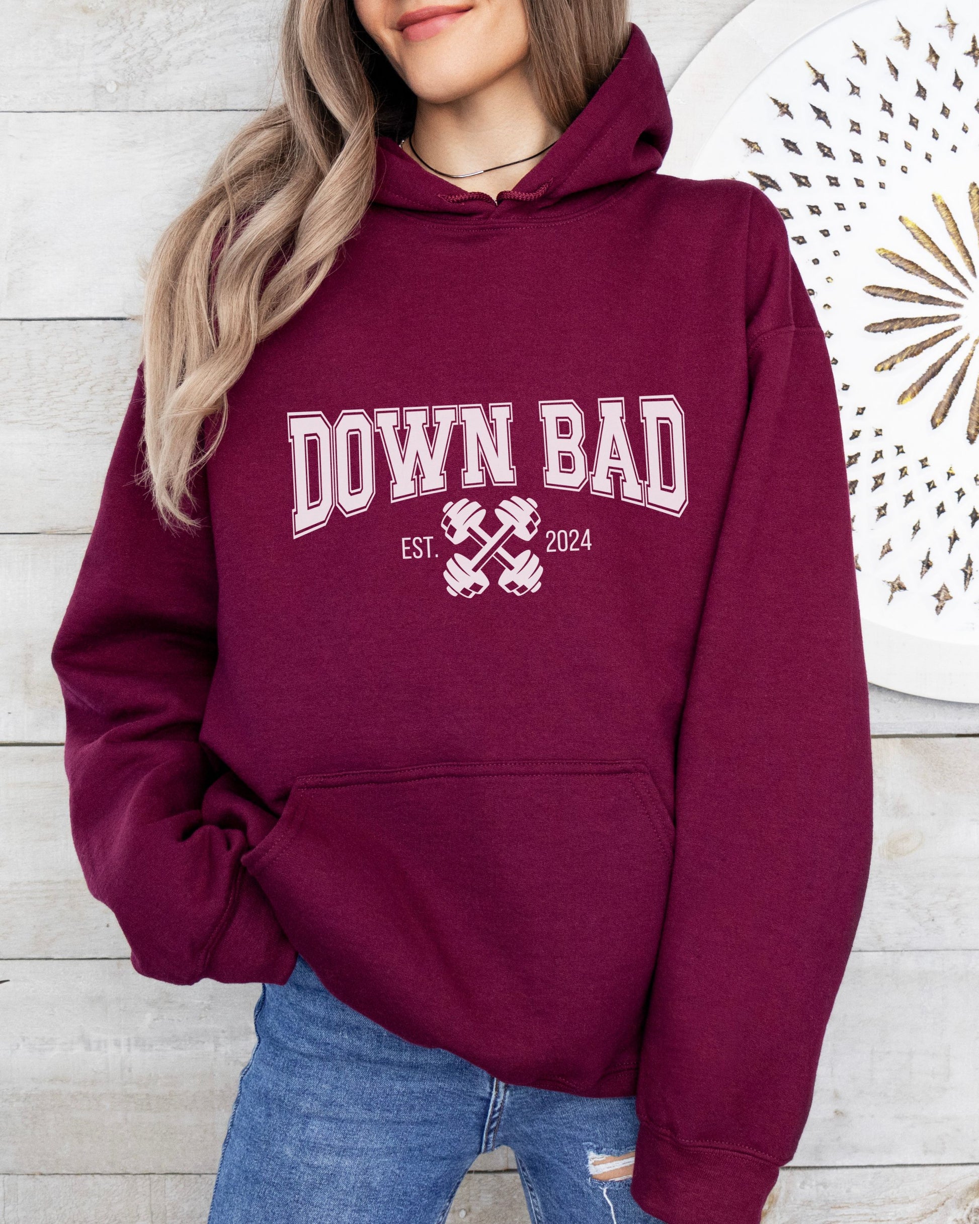 Maroon hoodie with 'DOWN BAD' text worn by a person against a wooden wall.