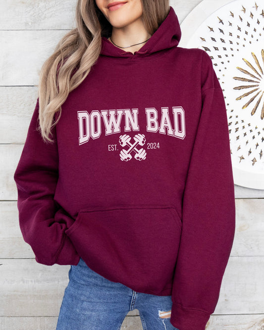 Maroon hoodie with 'DOWN BAD' text worn by a person against a wooden wall.