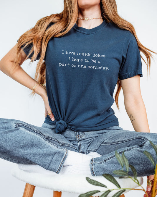 Person wearing a navy blue t-shirt with a humorous text design, sitting on a white stool.