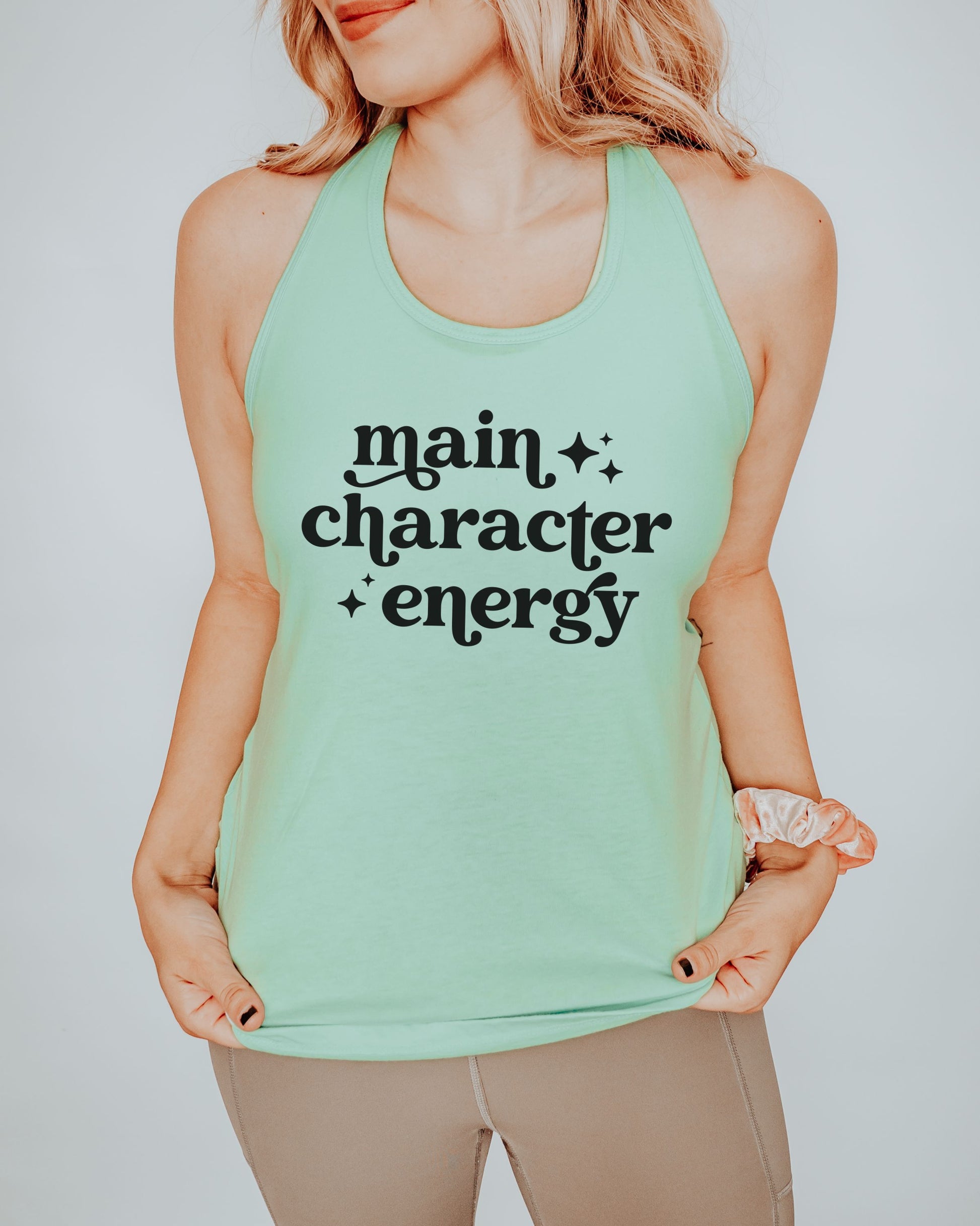 Person wearing a mint green tank top with 'main character energy' text on a plain background