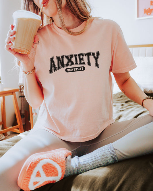 Person wearing a pink t-shirt with 'ANXIETY UNIVERSITY' text, holding a coffee cup.