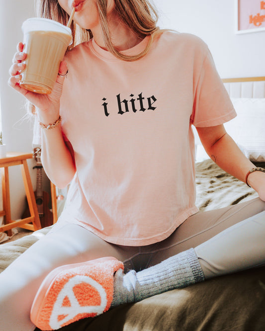 Person wearing a pink t-shirt with 'i bite' text, holding a coffee cup indoors.
