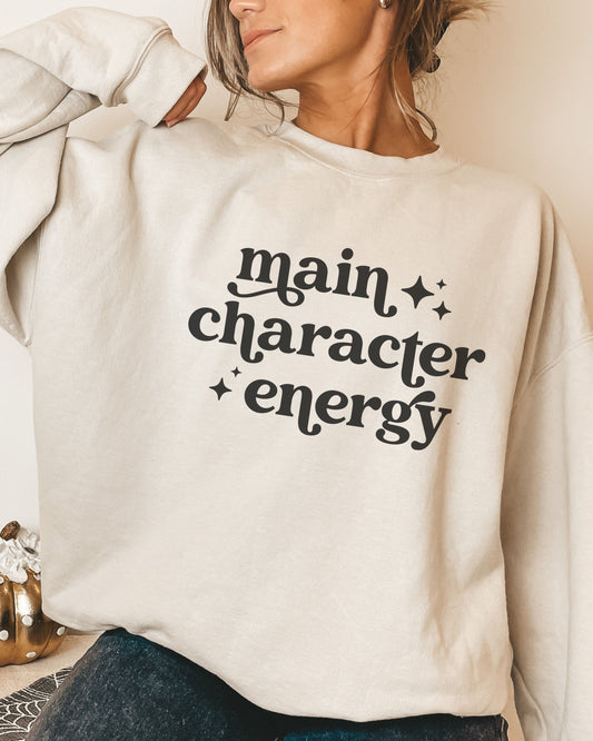 Person wearing a beige sweatshirt with 'main character energy' text.