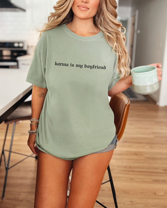 Person wearing a green t-shirt with 'karma is my boyfriend' text, holding a mug in a kitchen.