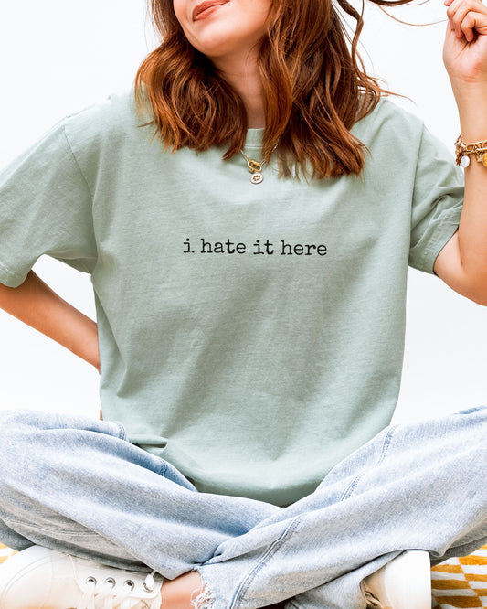 Person wearing a light green t-shirt with 'i hate it here' text.
