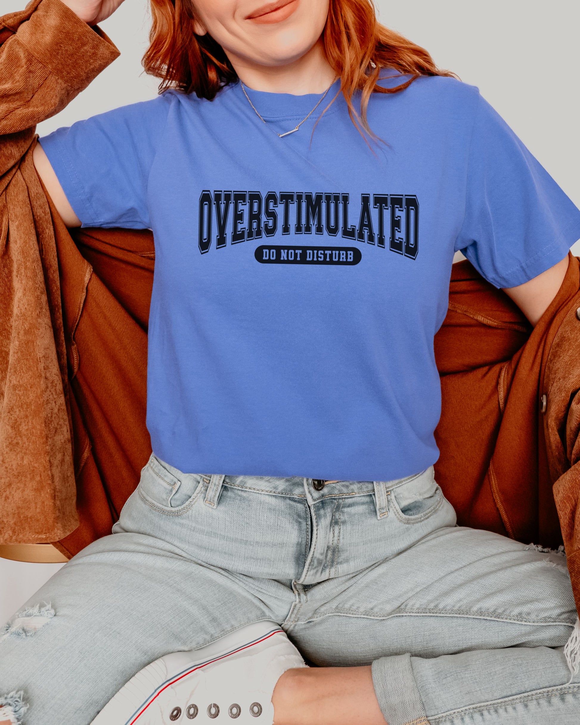 Person wearing a blue t-shirt with 'OVERSTIMULATED DO NOT DISTURB' text, sitting on a brown couch.