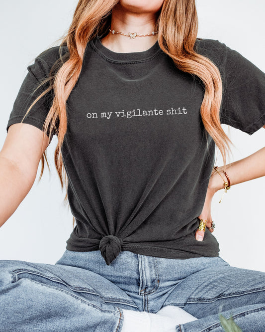 Person wearing a graphite t-shirt with on my vigilante shit text, sitting against a light background
