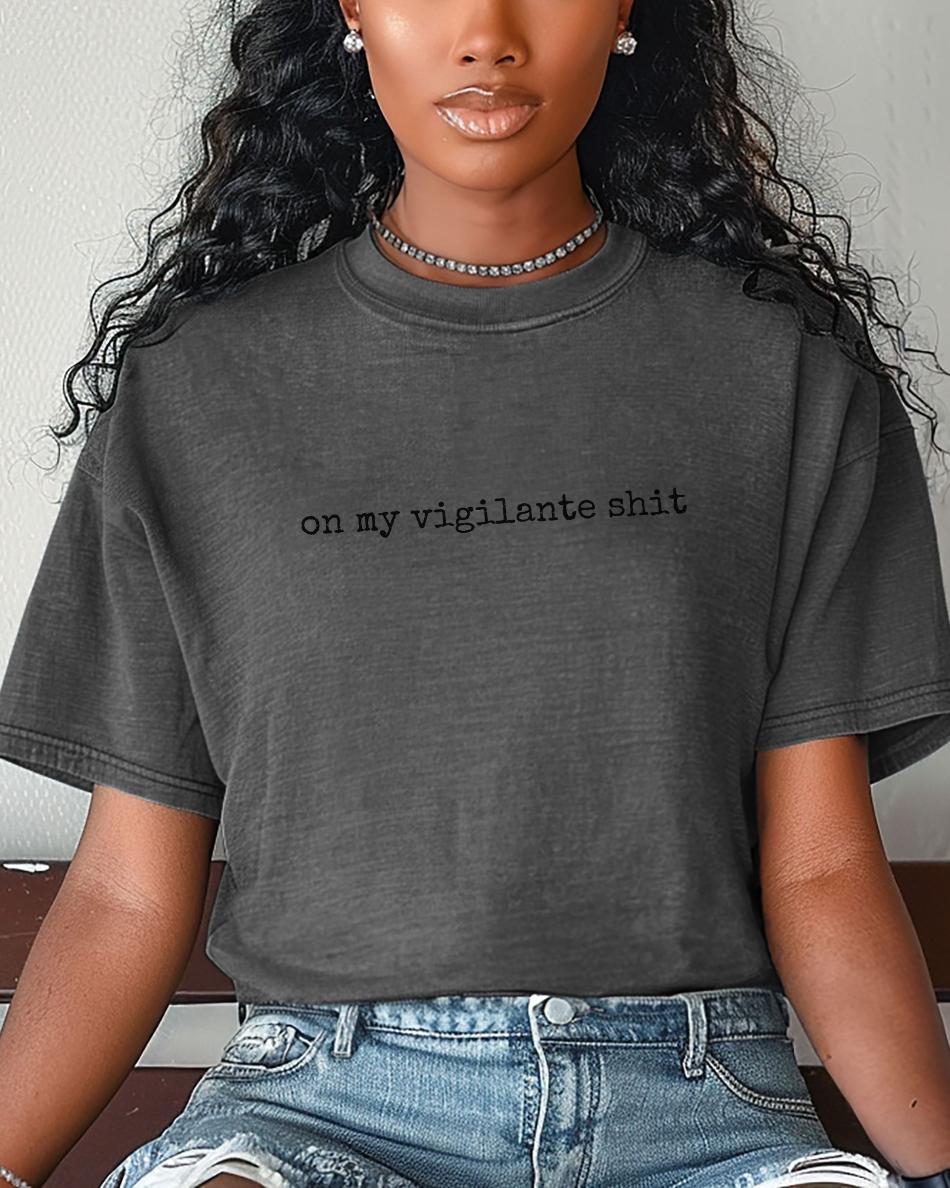 Person wearing a dark gray t-shirt with on my vigilante shit text with black font, sitting on a bench.